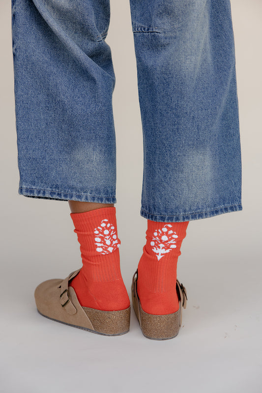 Red socks with white floral patterns worn with blue jeans and brown shoes on a light gray background