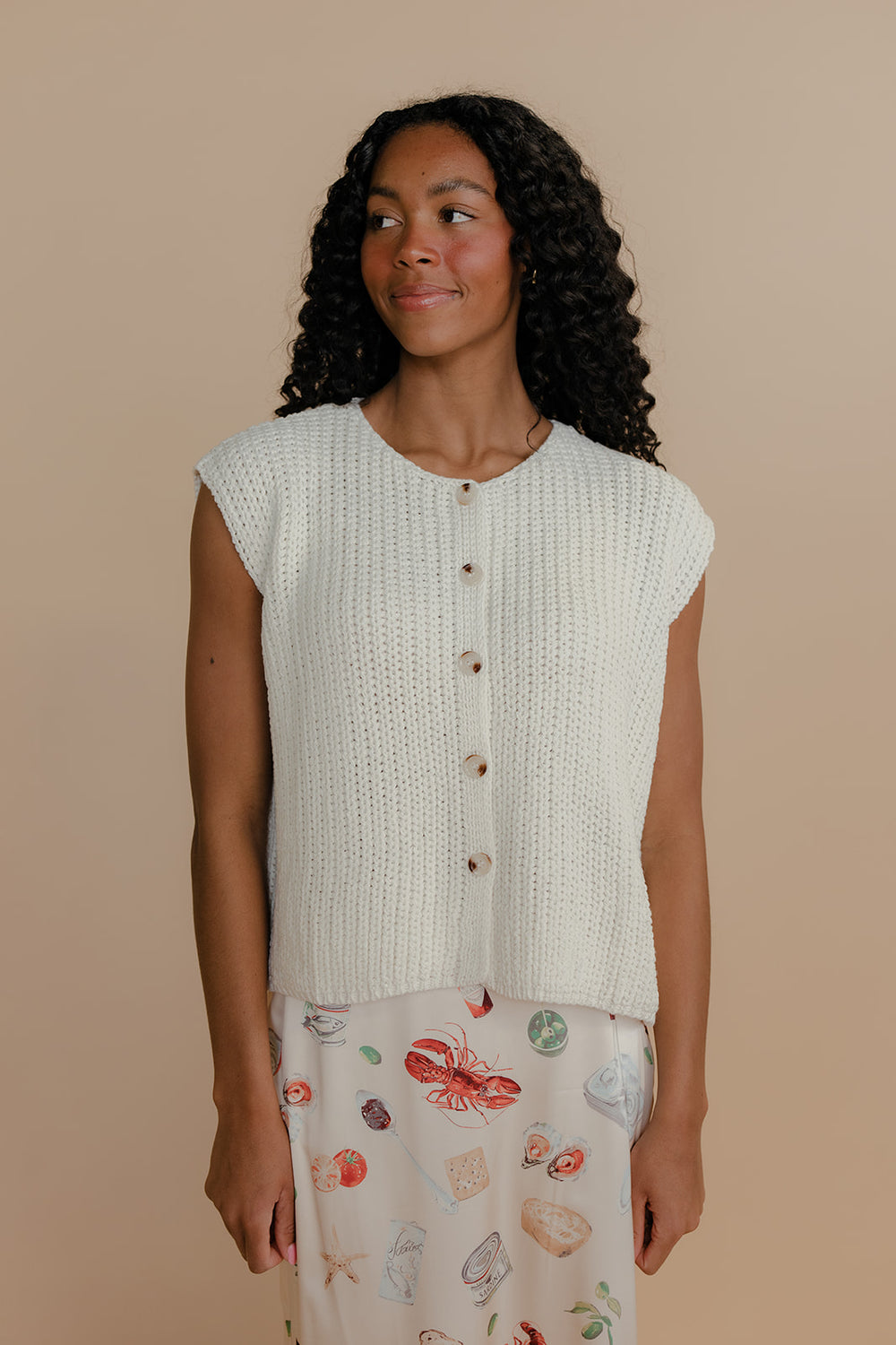 Woman wearing a light-colored knitted vest over a patterned skirt on a beige background