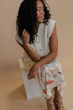 Woman sitting on a white stool wearing a patterned top and floral skirt against a beige background