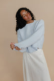Woman wearing a light blue sweater and white pants against a beige background