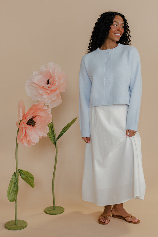 Woman in light blue top and white skirt standing next to large pink flowers on a beige background