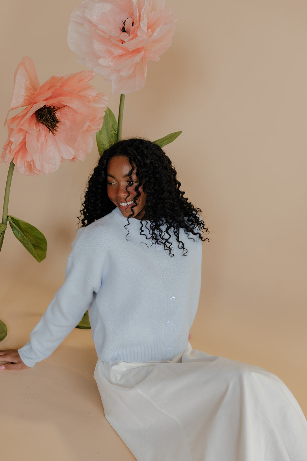 Woman in a light blue sweater sitting next to large pink flowers against a beige background