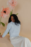 Woman in a light blue sweater sitting next to large pink flowers against a beige background