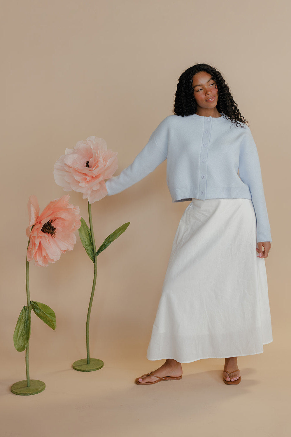 Woman in light blue top and white skirt standing next to large pink flowers on a beige background