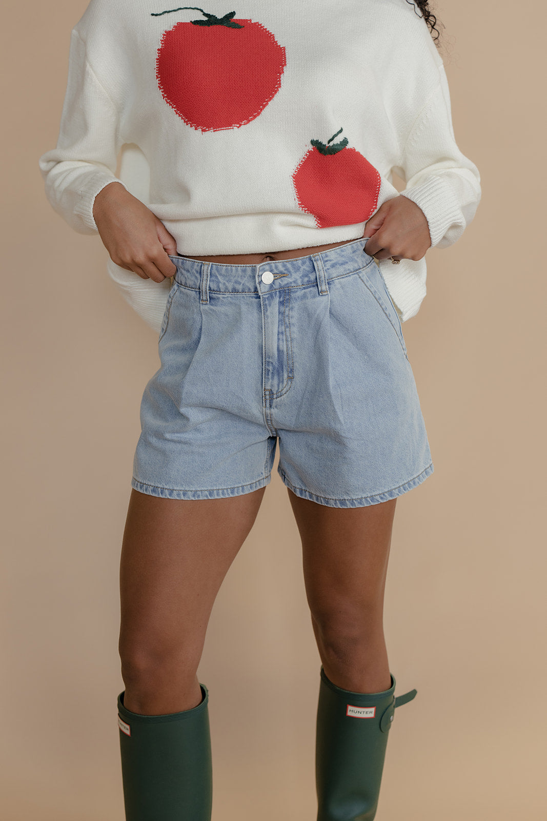 Person wearing a white sweater with red tomato designs, blue shorts, and green boots on a beige background