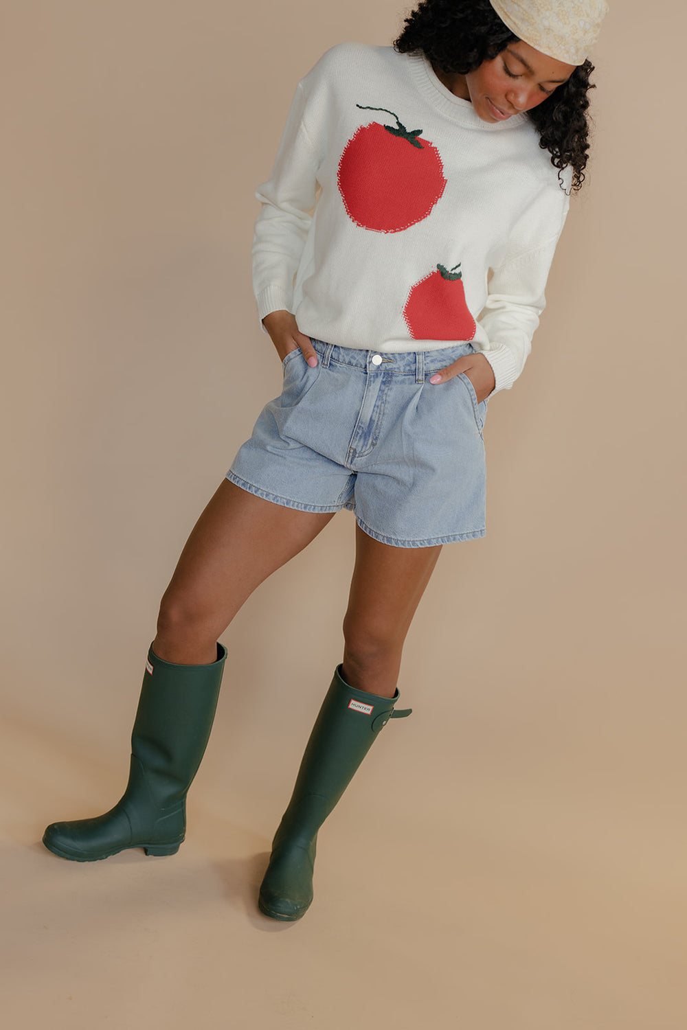 Person wearing a white sweater with red apple designs, denim shorts, and green rain boots on a beige background