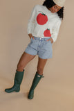 Person wearing a white sweater with red apple designs, denim shorts, and green rain boots on a beige background