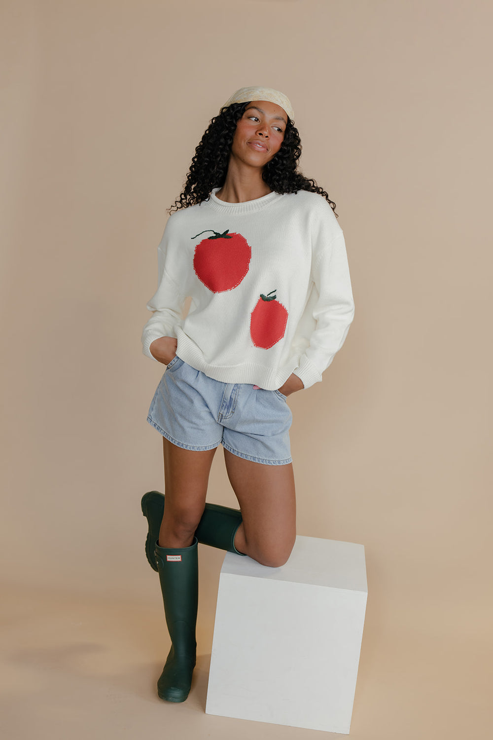 Person wearing a sweater with tomato designs, denim shorts, and green boots on a beige background