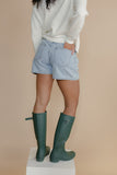Person wearing light blue denim shorts and green rain boots on a beige background