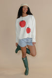 Woman wearing a sweater with red apple designs, denim shorts, and green rain boots on a beige background