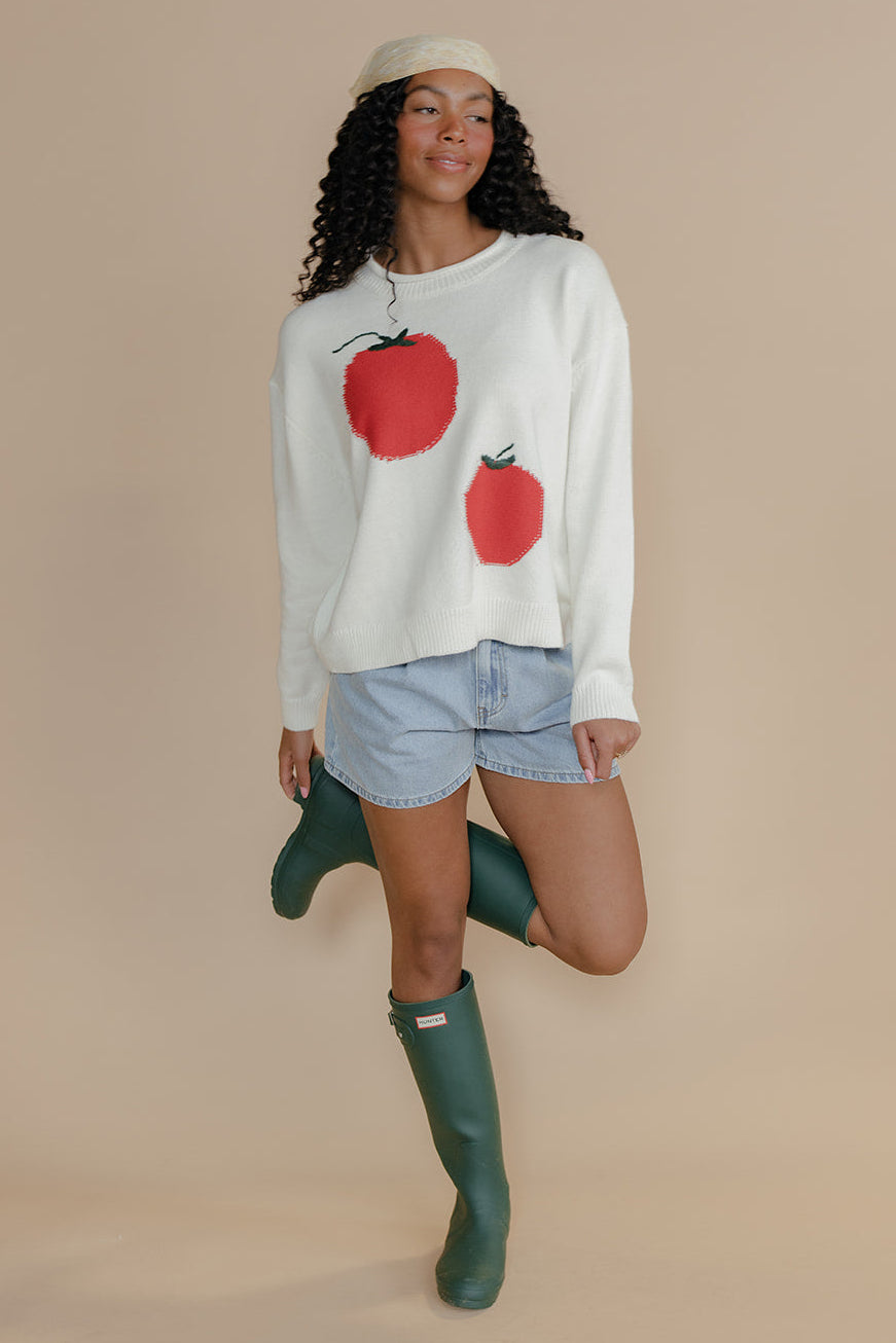 Woman wearing a sweater with red apple designs, denim shorts, and green rain boots on a beige background