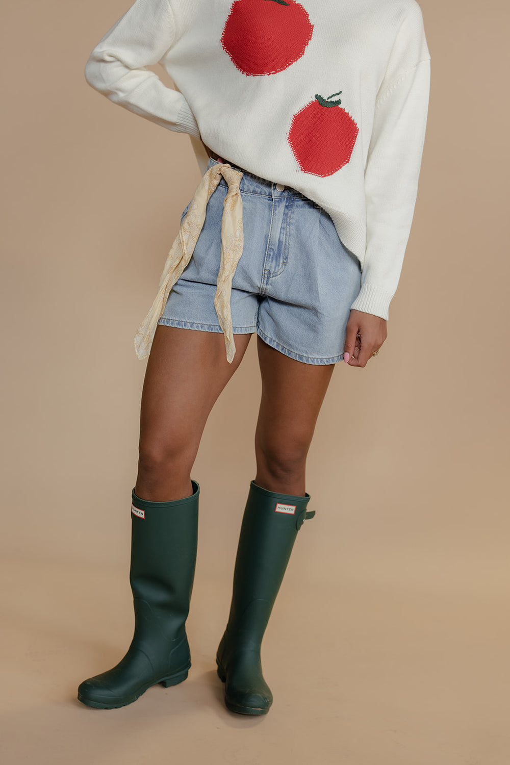 Person wearing a white sweater with red apples, denim shorts, and green rain boots on a beige background