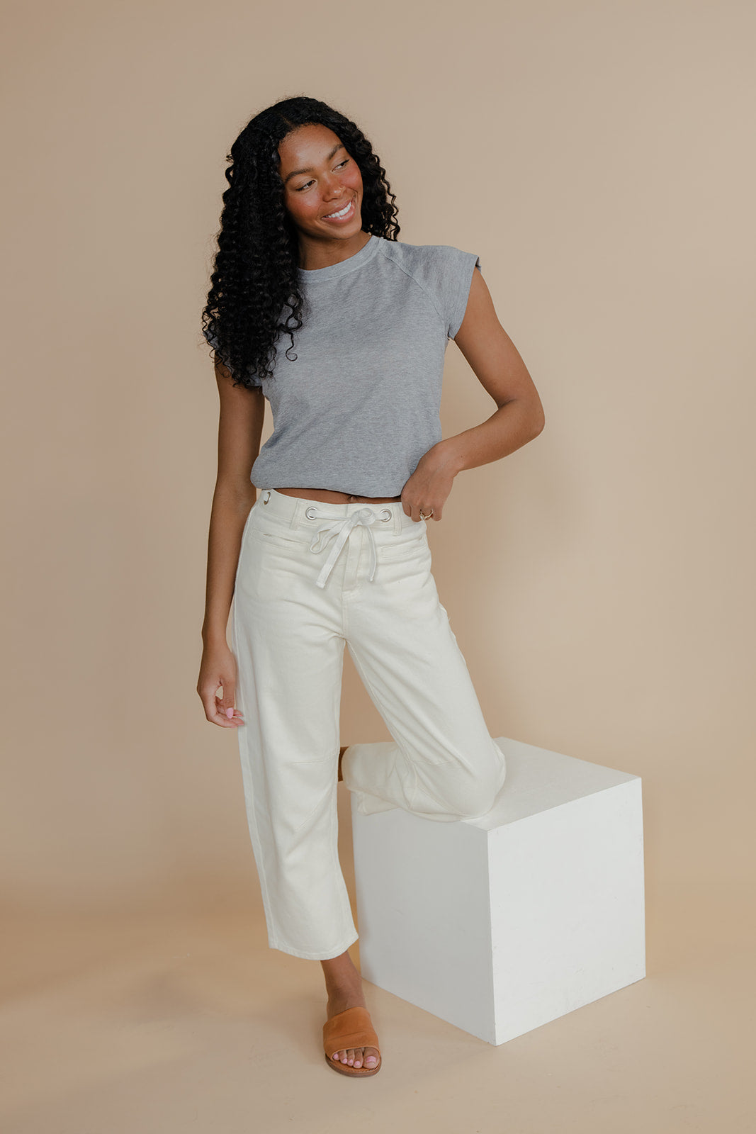 Woman wearing a gray sleeveless top and white pants standing on a white block against a beige background
