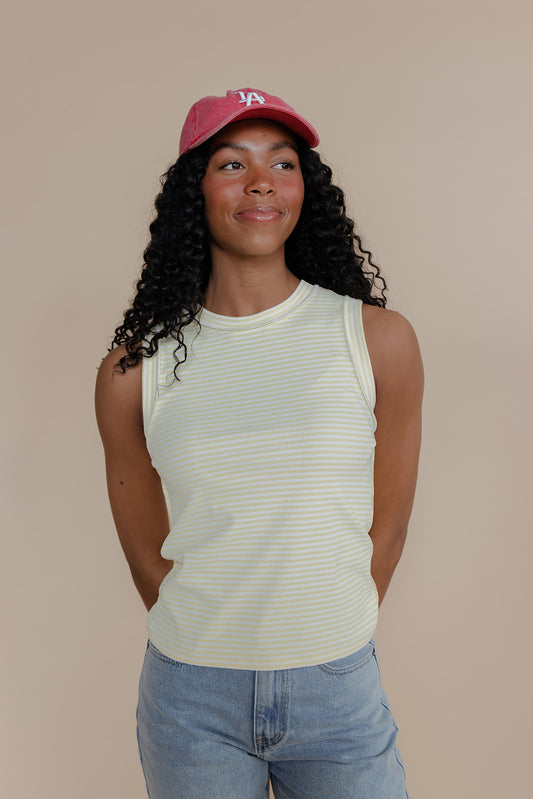 Woman wearing a red cap, white striped tank top, and blue jeans against a beige background