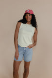 Woman wearing a light green sleeveless top and denim shorts with a pink cap against a beige background