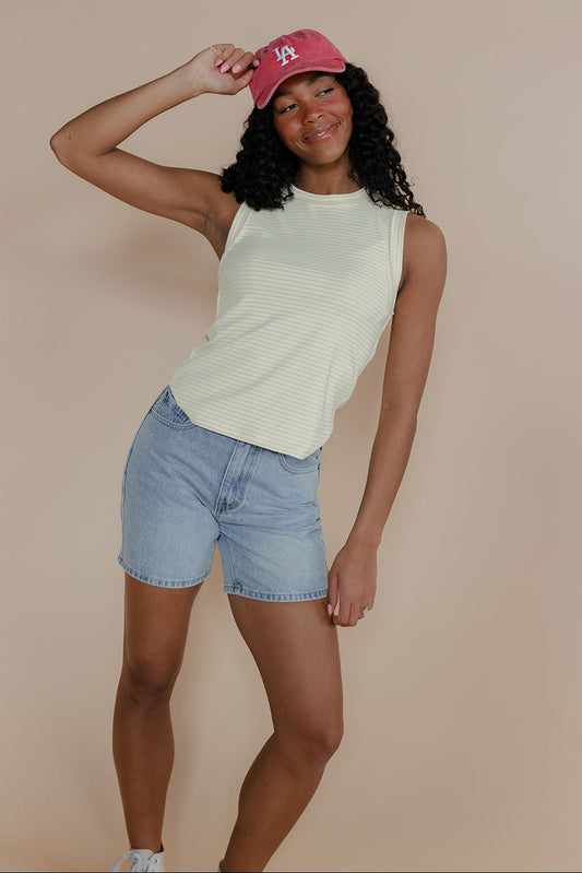 Woman wearing a light green sleeveless top and denim shorts, posing against a beige background.