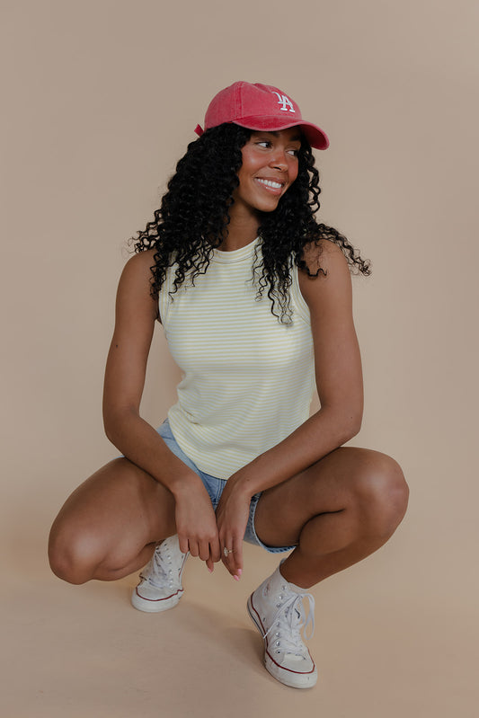 Woman wearing a red cap, white tank top, denim shorts, and white sneakers on a beige background