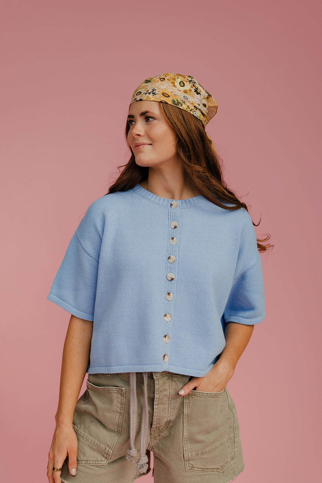 Woman wearing a light blue shirt with buttons, green shorts, and a patterned headscarf on a pink background