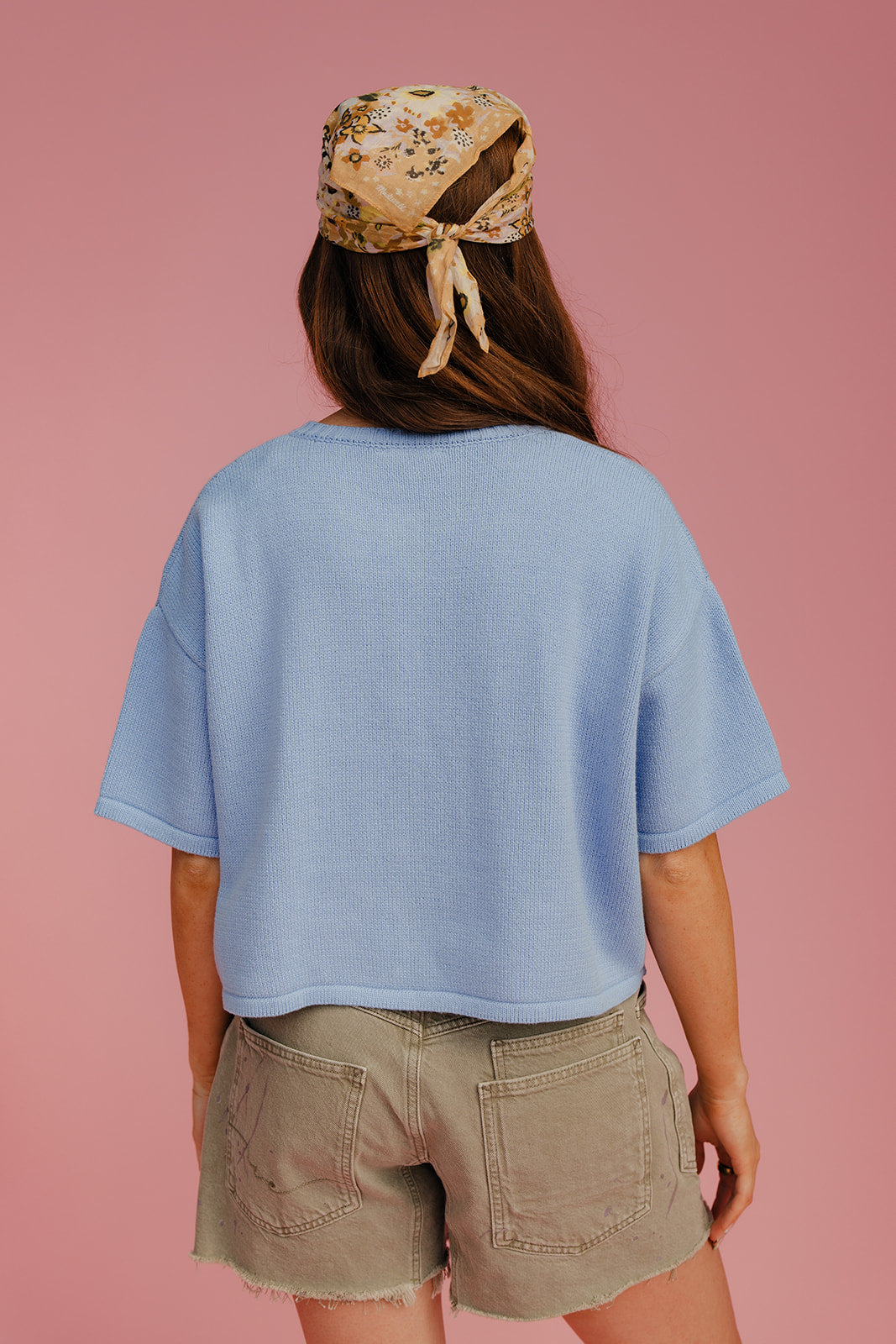 Person wearing a light blue shirt and beige shorts with a floral headscarf on a pink background