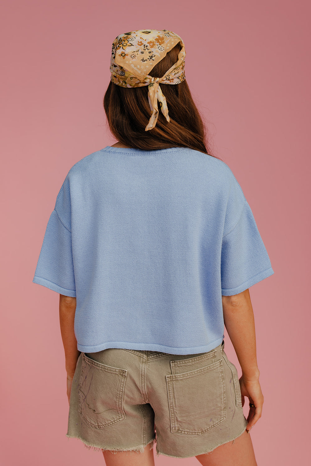 Person wearing a light blue shirt and beige shorts with a floral headscarf on a pink background