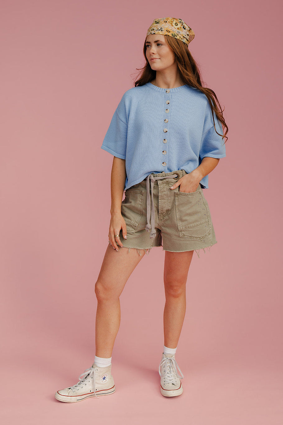 Woman wearing a light blue shirt, beige shorts, and white sneakers on a pink background