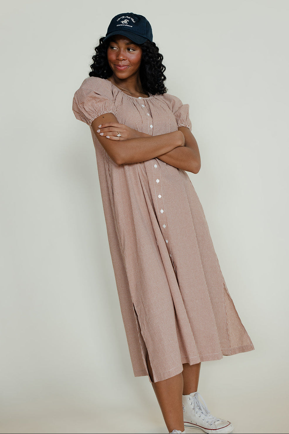 Woman wearing a long beige dress and navy cap against a white background