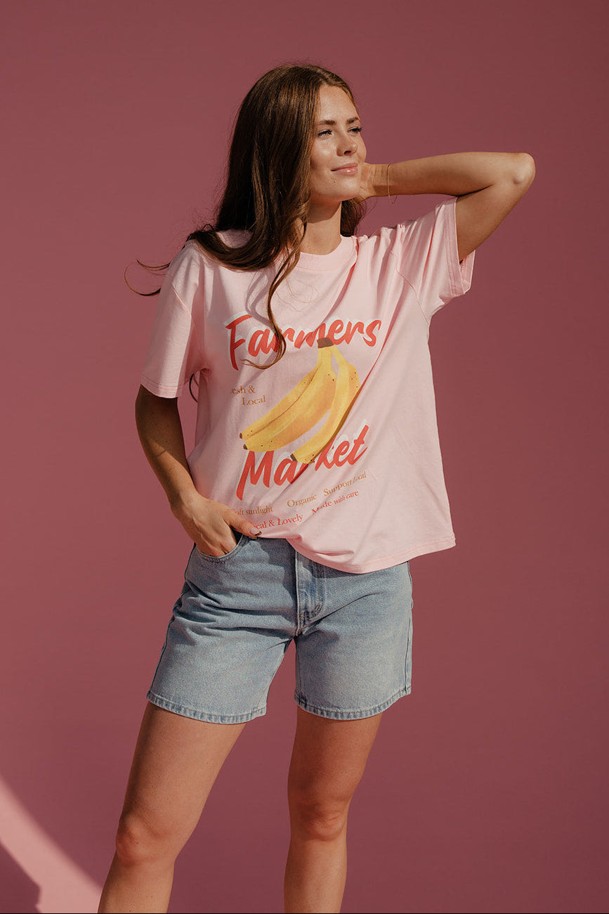 Woman wearing a pink t-shirt with 'Farmers Market' and banana graphic on a pink background