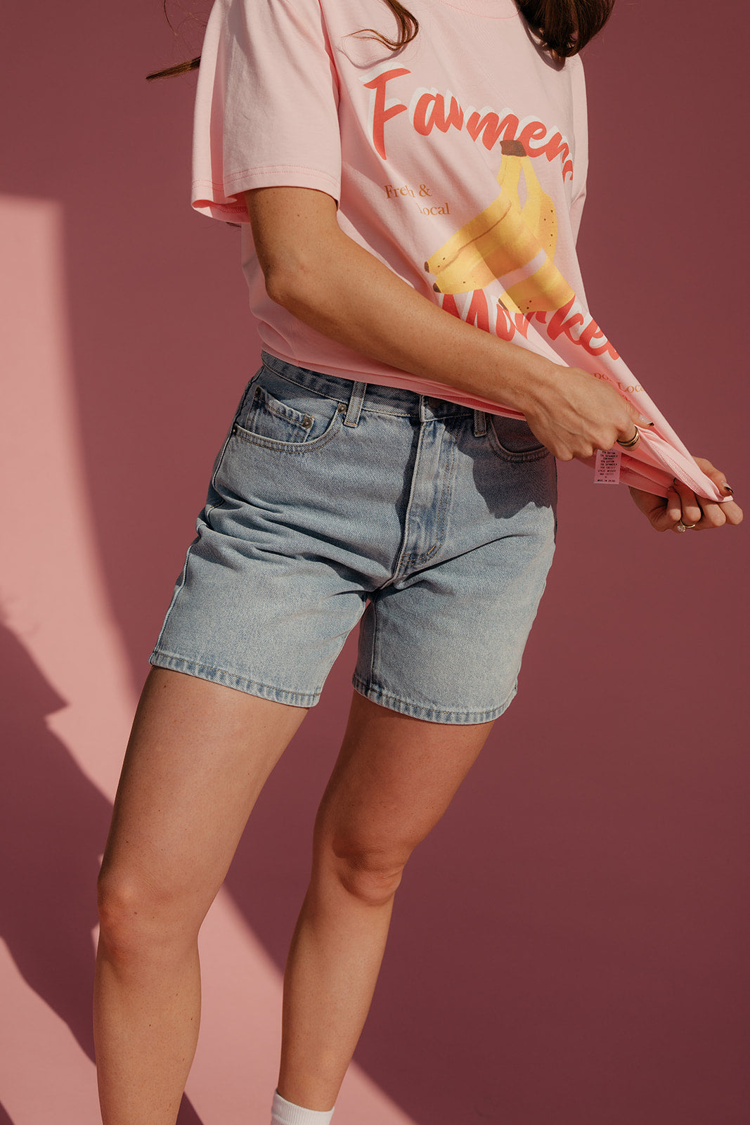Person wearing a pink t-shirt with a graphic design and denim shorts against a pink background