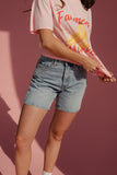 Person wearing a pink t-shirt with a graphic design and denim shorts against a pink background