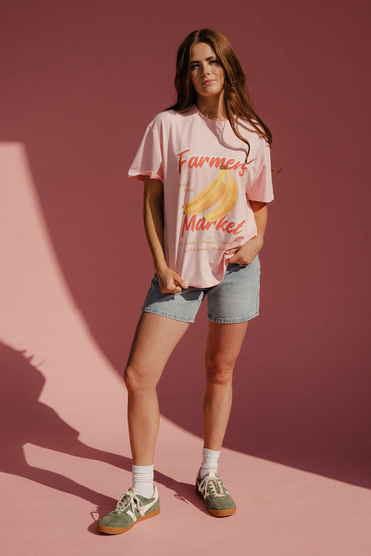 Woman wearing a pink t-shirt with 'Farmers Market' and banana graphic against a pink background