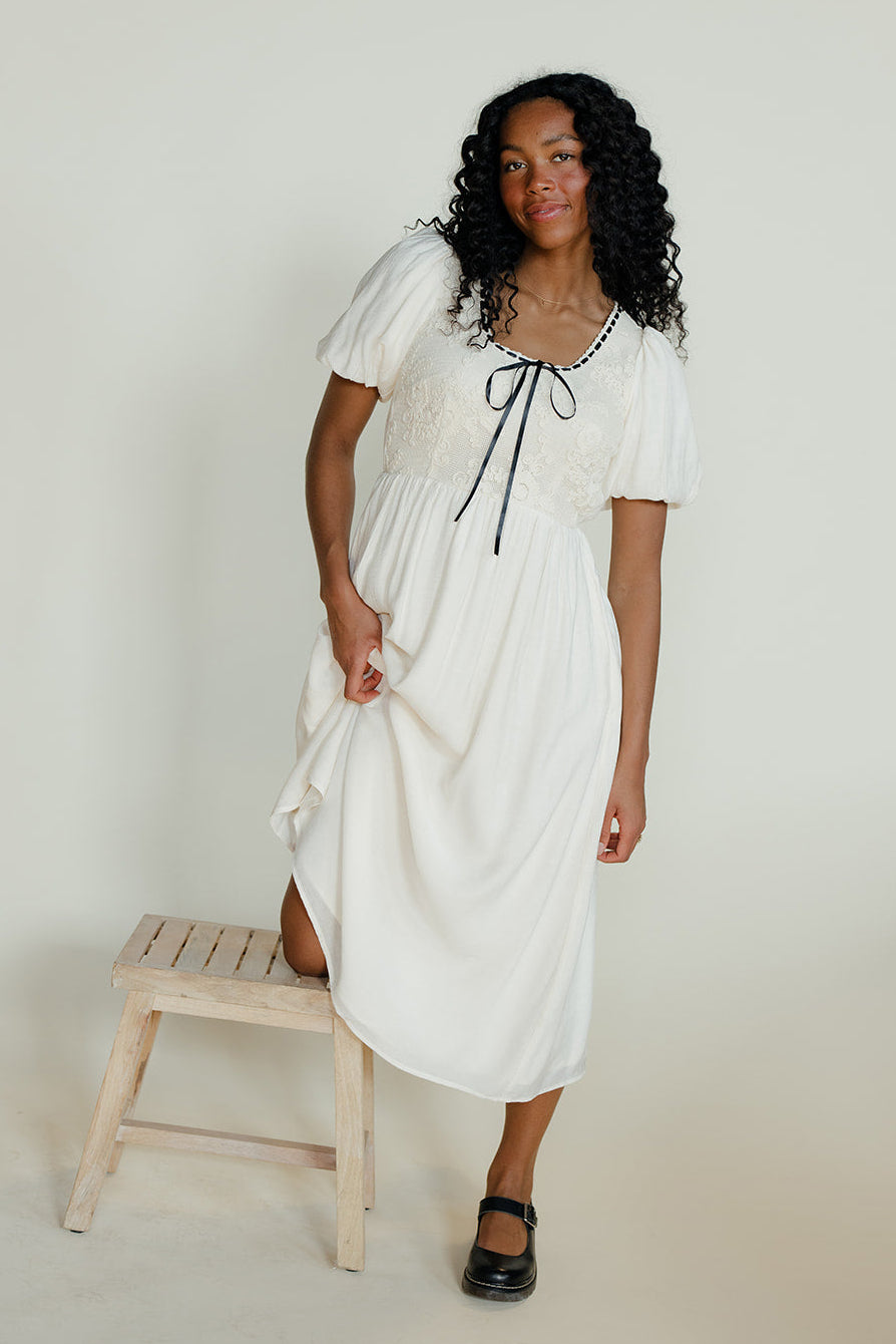 Woman in a white dress standing next to a wooden stool against a plain background