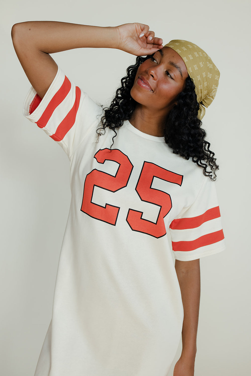 Woman wearing a white t-shirt with red numbers and a beige cap against a plain background