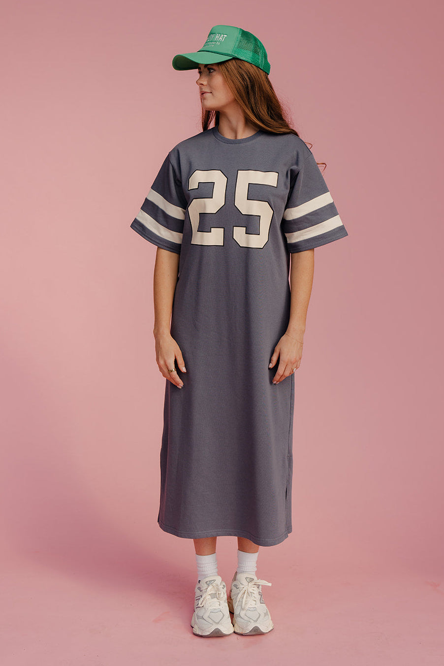 Woman wearing a gray dress with number 25 and a green cap on a pink background