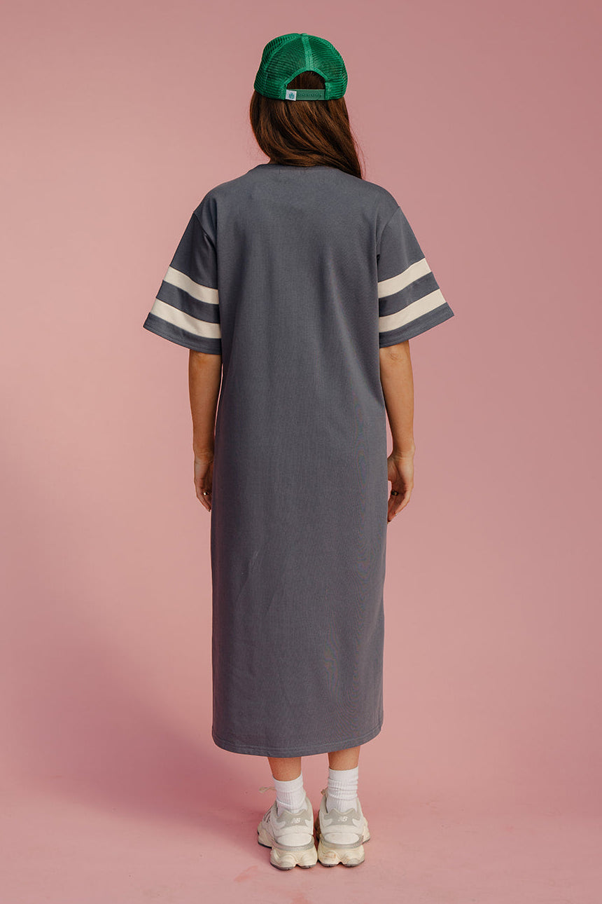 Person wearing a long gray dress with white stripes and a green cap on a pink background