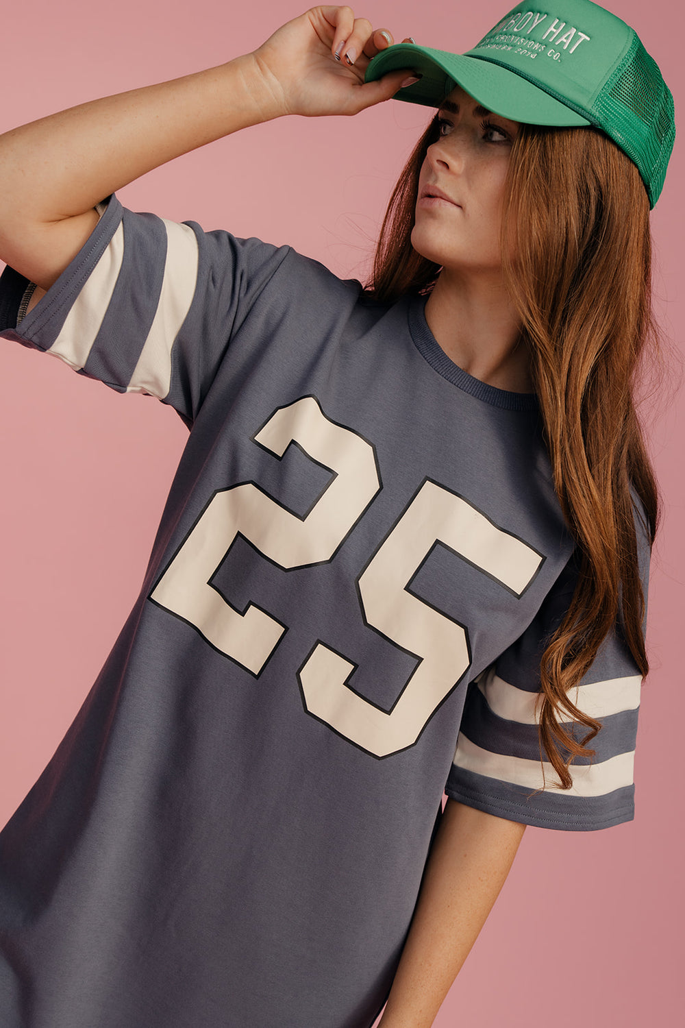 Person wearing a gray t-shirt with the number 25 and a green cap on a pink background