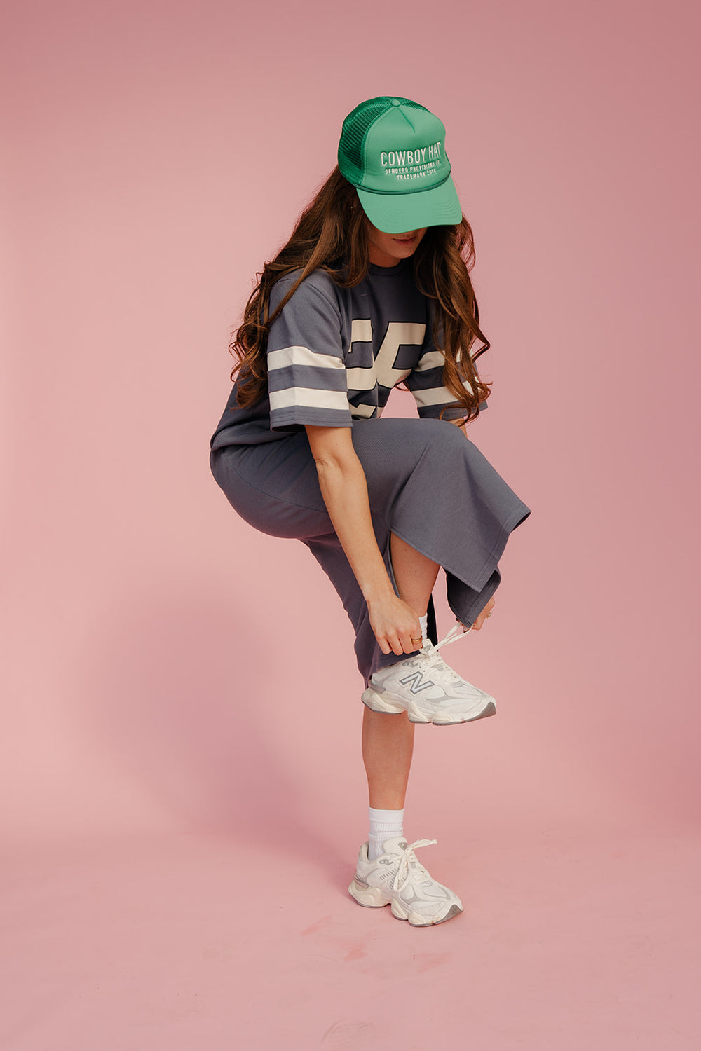 Person wearing a green cap, gray outfit, and white sneakers on a pink background