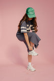 Person wearing a green cap, gray outfit, and white sneakers on a pink background
