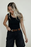 Woman wearing a black sleeveless top and black pants with a brown belt against a plain background