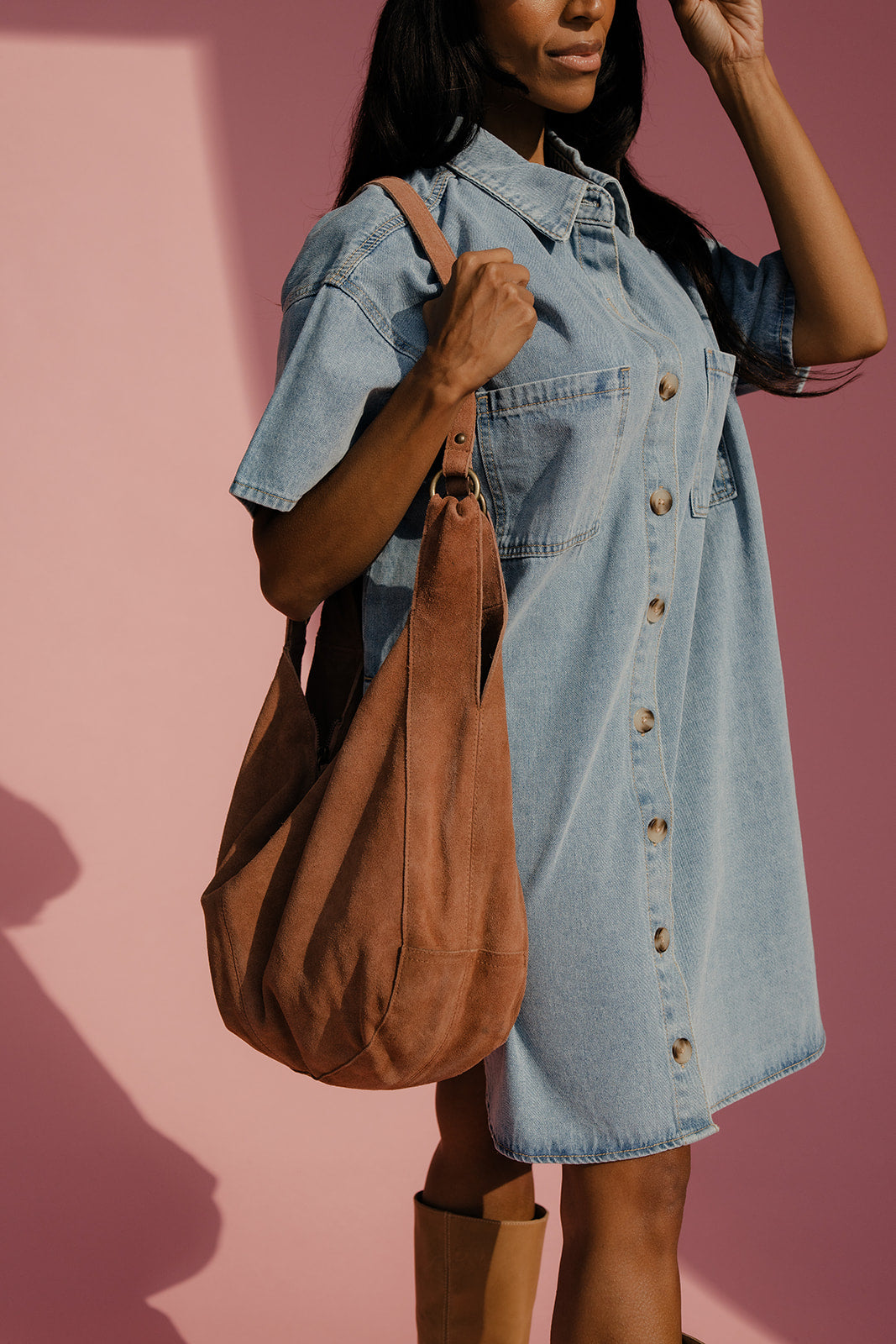 Woman wearing a denim dress holding a brown leather bag against a pink background