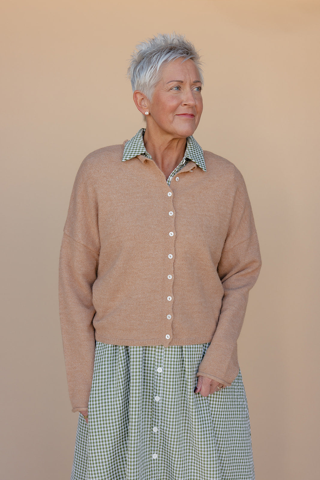Woman wearing a brown cardigan over a green checkered shirt and skirt against a beige background