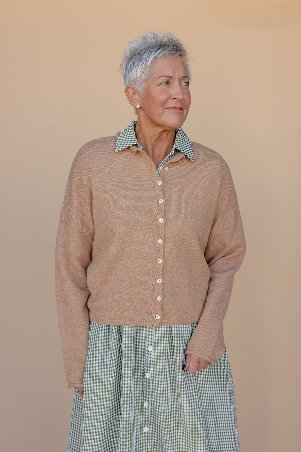 Woman wearing a brown cardigan over a green checkered shirt and skirt against a beige background