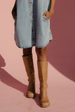 Woman wearing a denim dress and tan boots against a pink background
