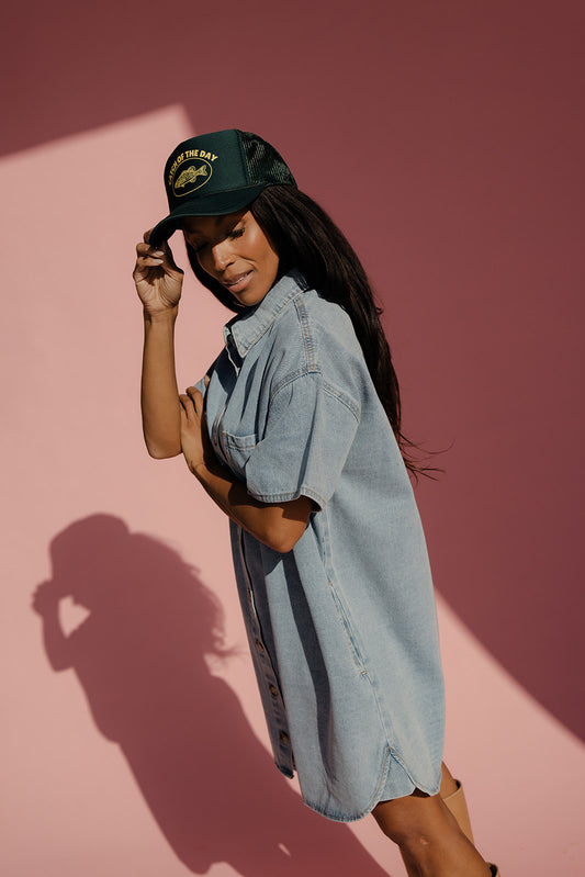 Woman wearing a denim dress and cap against a pink background