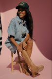 Woman wearing a denim jacket, cap, and knee-high boots sitting on a stool against a pink backdrop.
