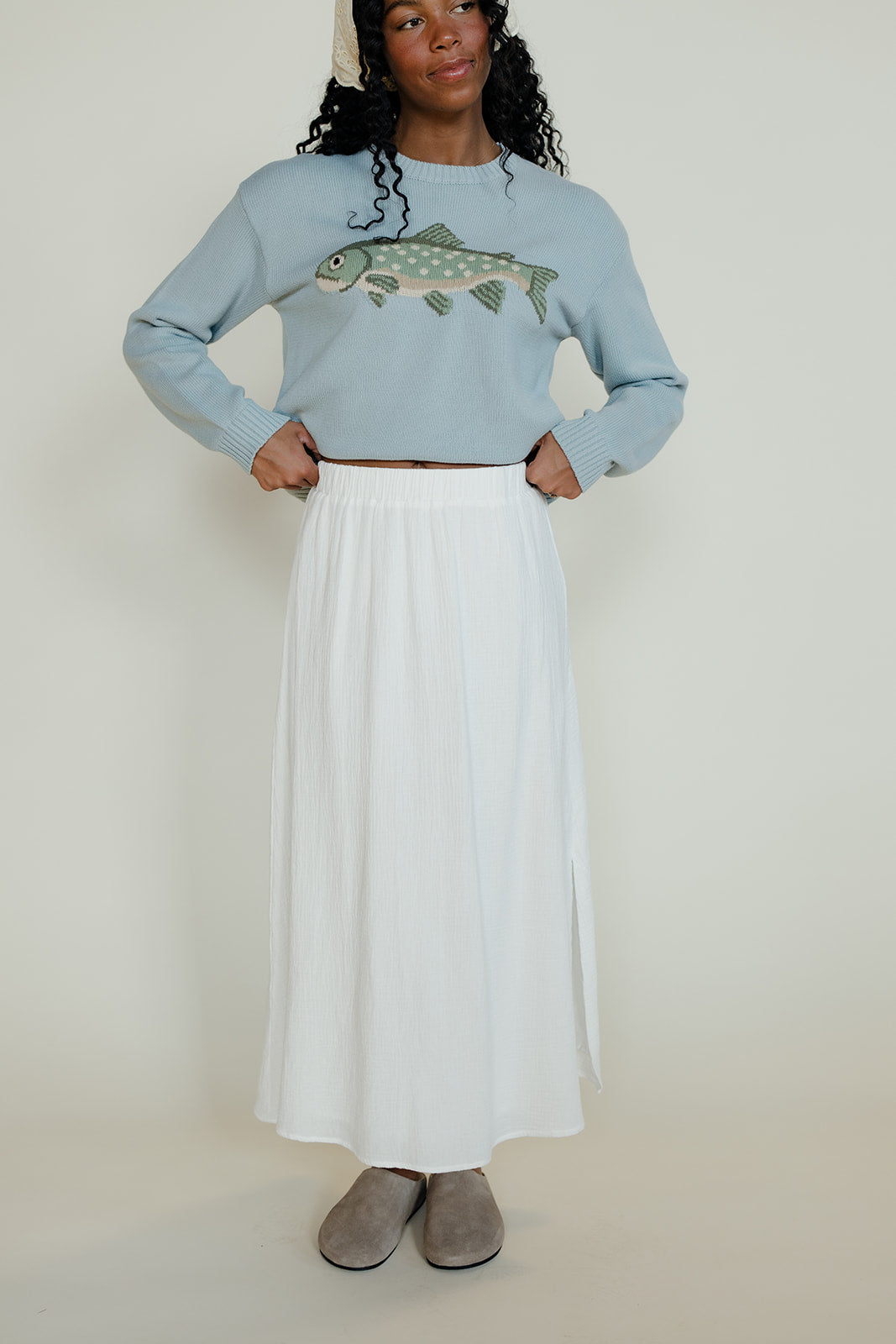 Person wearing a light blue sweater with a fish design and a white skirt on a plain background