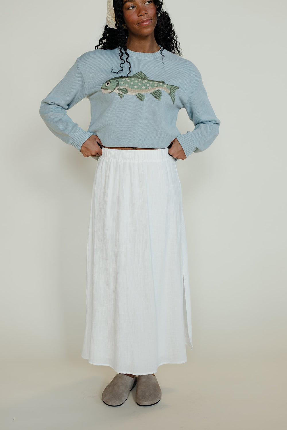 Person wearing a light blue sweater with a fish design and a white skirt on a plain background
