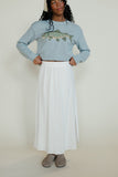 Person wearing a light blue sweater with a fish design and a white skirt on a plain background