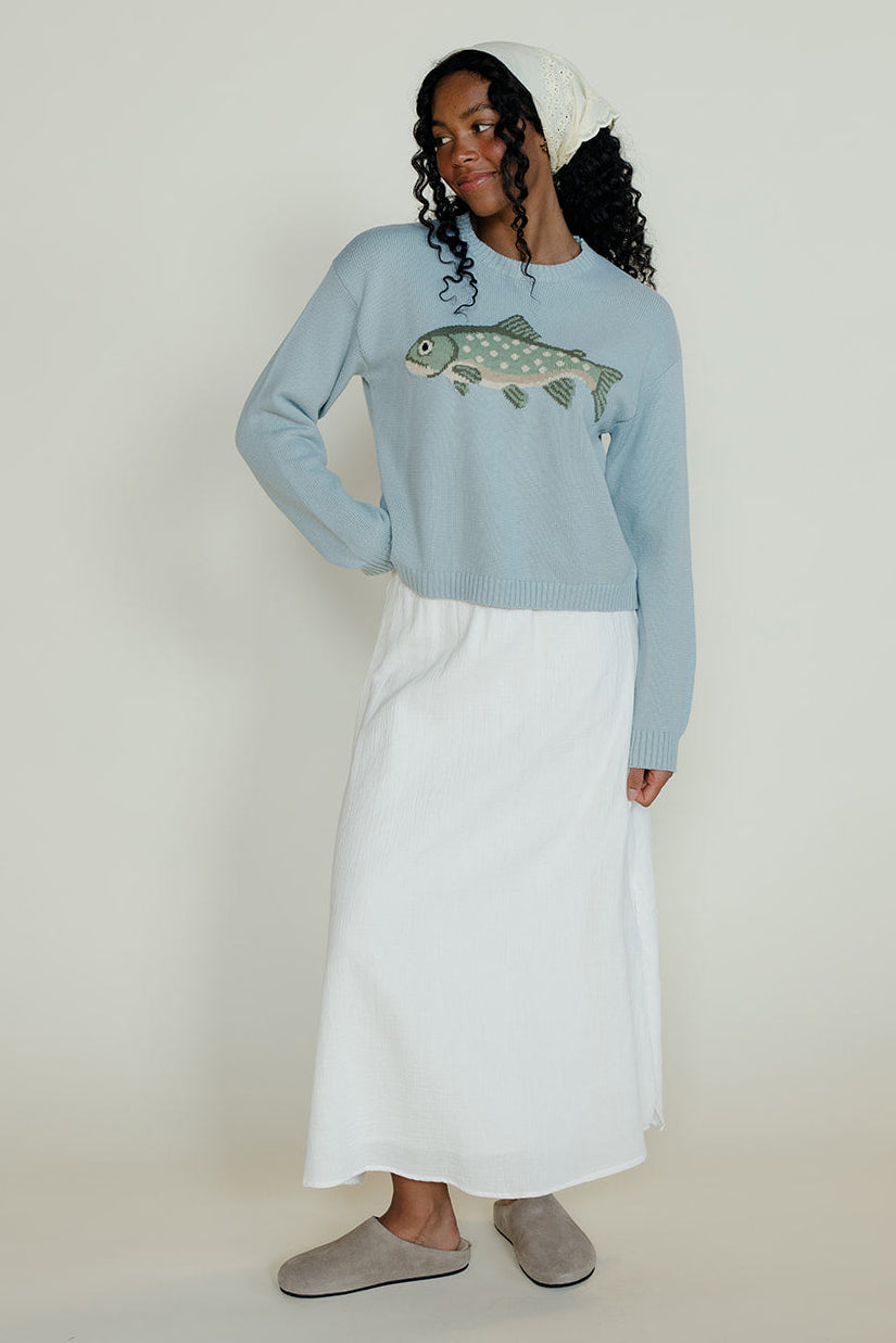 Woman wearing a light blue sweater with a fish design, white pants, and a white beanie on a plain background