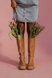 Person wearing brown knee-high boots with pink tulips peeking out, against a pink background
