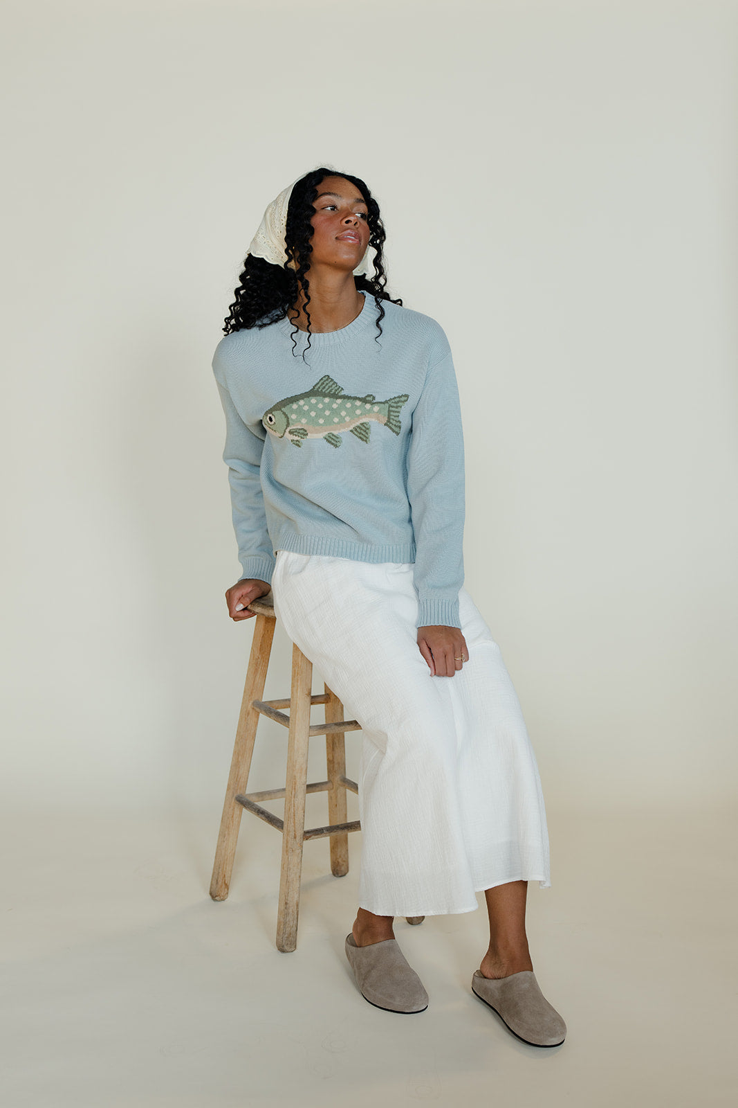 Woman wearing a light blue sweater with a fish design, standing on a stool against a plain background.